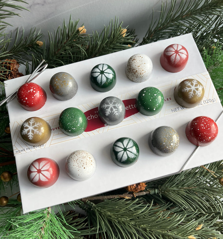 Assorted Christmas-themed chocolate truffles on a white card with greenery around. The Chocolate Palette Salt Lake City Utah